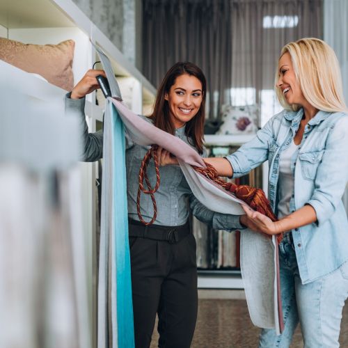 Young woman designer talking with a saleswoman and chooses fabrics for curtains in textile show room.